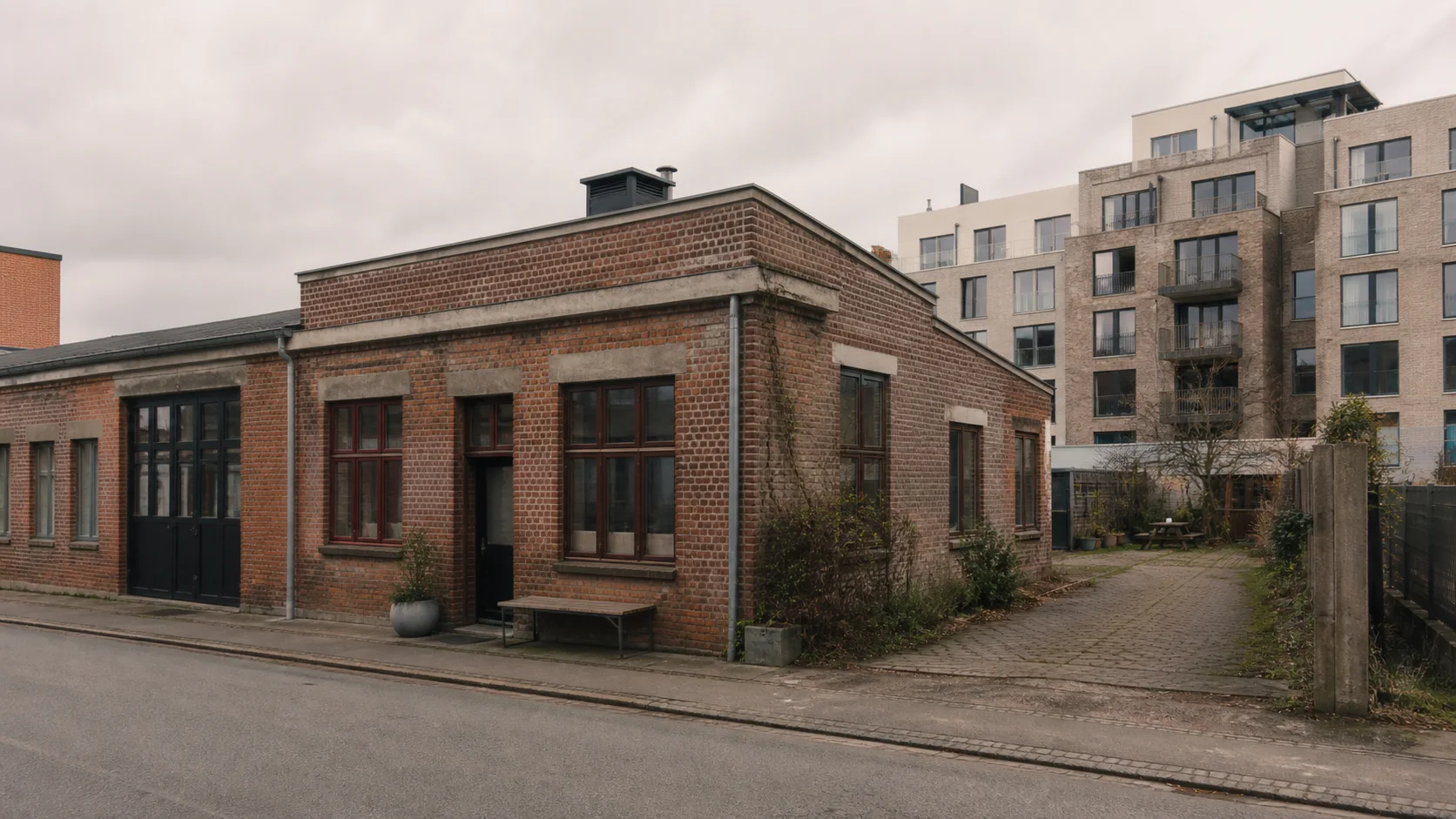 Lav ældre værkstedsbygning i mursten foran nyere københavnsk boligbyggeri og et lille underudnyttet gårdareal.