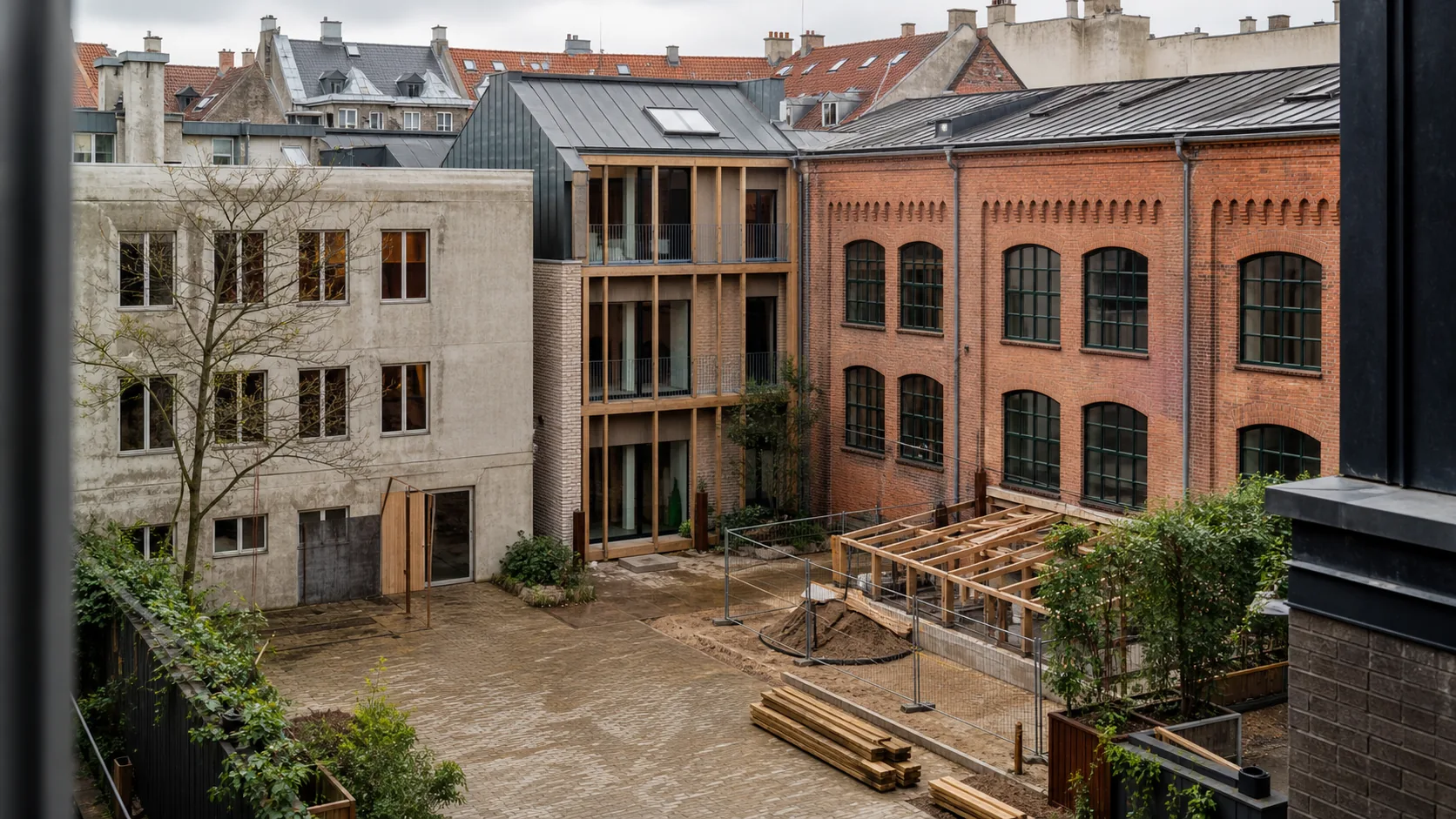 Københavnsk gårdrum med en ældre værkstedsbygning i rød mursten, en betonbygning og en nyere træ- og murstensbygning omkring en mindre byggeplads.