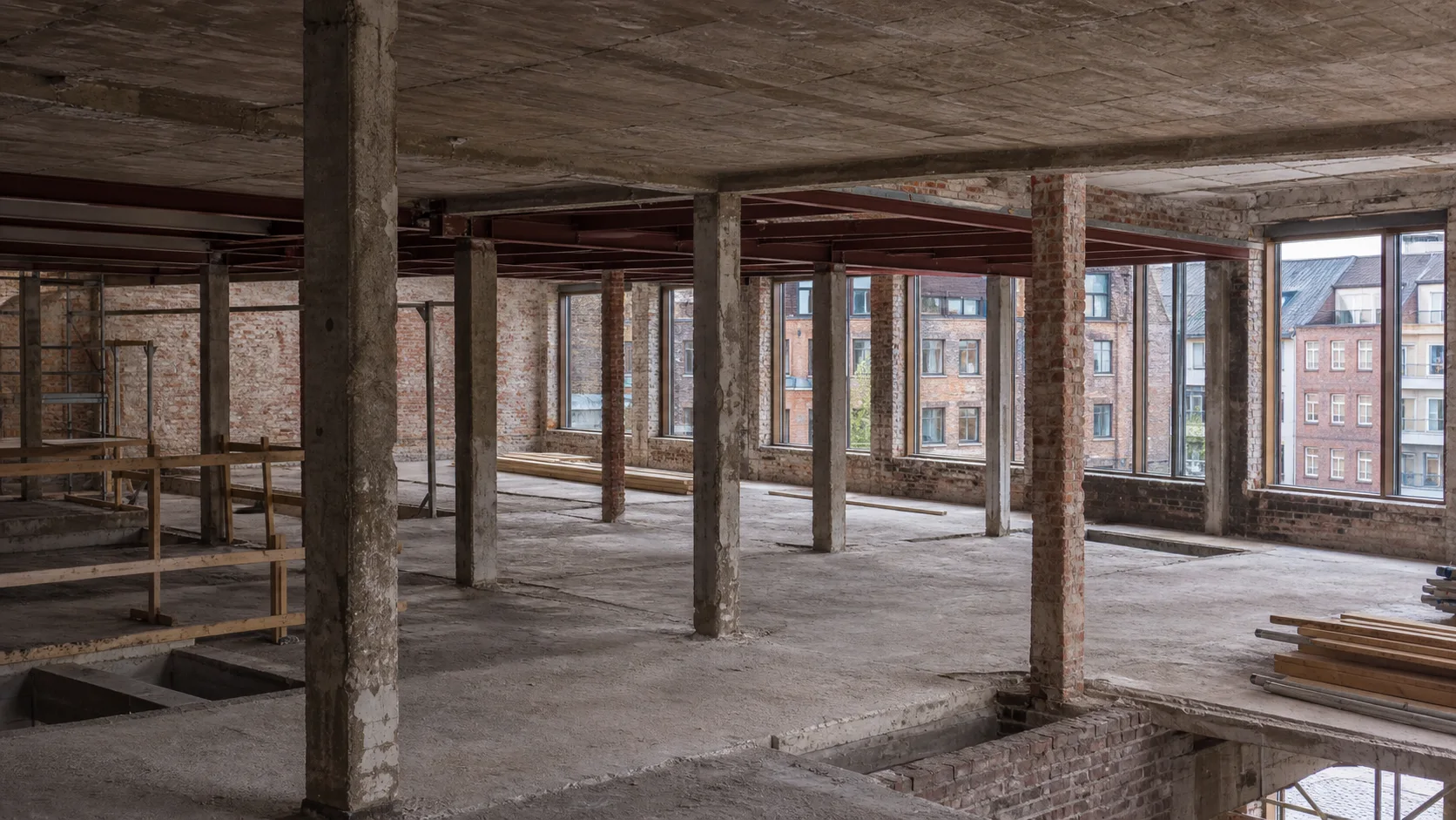 Indre af ældre erhvervsbygning under ombygning med synlige betonsøjler, murværk, stålbjælker og store vinduesåbninger.